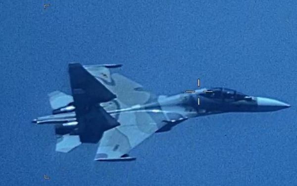 The image shows a military fighter jet photographed from above against a blue sky background. The aircraft displays a camouflage paint scheme and appears to be carrying underwing ordnance or external stores. The image contains targeting reticle overlays suggesting it was captured by a surveillance or targeting camera system.