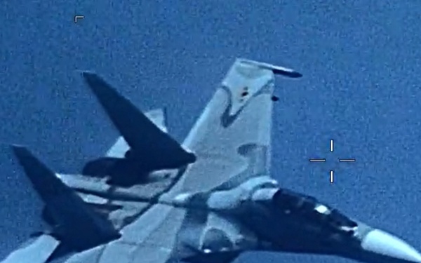 An aerial surveillance or targeting camera image showing a military fighter jet photographed from above or nearby aircraft. The image has the characteristic appearance of an electro-optical or infrared targeting pod feed, indicated by the crosshair/reticle overlay visible on the right side. The aircraft appears to be in flight against a blue sky background.