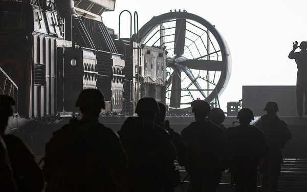 A group of military personnel in silhouette are moving across what appears to be the well deck of a naval amphibious assault ship, likely boarding or disembarking from a Landing Craft Air Cushion (LCAC) hovercraft. One individual at the far end appears to be directing or signaling with raised hands. The image is strongly backlit, creating dramatic silhouettes against the bright exterior light.