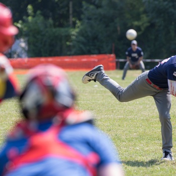 A baseball or softball game is in progress outdoors on a grass field. A pitcher in a navy blue jersey and gray pants has just released a pitch, with the ball visible in the air. In the foreground, a batter in red and a catcher in red/blue gear are visible but out of focus.