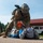 Paralegal training ensures disciplined forces are lethal forces A U.S. Army soldier in full combat gear and ACU/OCP camouflage uniform is conducting a detainee control or apprehension drill on a paved surface. A role-player wearing a keffiyeh scarf is positioned face-down on the ground while the soldier applies a restraint technique. A safety observer in an orange vest is visible in the background, indicating this is a training exercise.