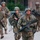 Paralegal training ensures disciplined forces are lethal forces A group of military soldiers in full combat gear are moving in formation along a paved path. The lead soldier is in the foreground wearing MultiCam (OCP) pattern uniform, carrying an M16-style rifle, with a combat helmet and protective eyewear. Additional soldiers in the background wear ACU (Army Combat Uniform) pattern, suggesting a mixed unit during a training exercise or movement drill.
