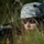 Paralegal training ensures disciplined forces are lethal forces A soldier in U.S. Army ACU (Army Combat Uniform) digital camouflage is lying prone in tall grass, partially concealed by vegetation. The individual is wearing a MICH/ACH combat helmet and ballistic eyewear while holding what appears to be a rifle. The scene suggests a field training exercise or tactical movement drill.
