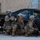 Paralegal training ensures disciplined forces are lethal forces Four U.S. military personnel in full combat gear are taking cover behind a dark-colored sedan with shattered windows. They are in a crouched tactical position, armed with rifles, adjacent to a building with a tan brick exterior. The vehicle's windows appear to be broken, suggesting a training exercise or active incident scenario.