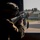 Paralegal training ensures disciplined forces are lethal forces A military service member in full combat gear is aiming a rifle through a window or opening toward an outdoor area. The soldier is wearing a combat helmet with attachments and a uniform bearing a US flag patch on the sleeve. The setting appears to be a military installation or training facility based on the visible infrastructure in the background.