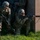 Paralegal training ensures disciplined forces are lethal forces Two U.S. Army soldiers in full combat gear are conducting a military training exercise near the corner of a cinder block building. One soldier is kneeling and aiming an M16-style rifle around the corner while the other stands in a ready position. The rifles are equipped with red MILES (Multiple Integrated Laser Engagement System) training devices, confirming this is a simulated combat exercise.