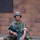 Paralegal training ensures disciplined forces are lethal forces A soldier in full military uniform sits on a white stool in front of a brick wall, holding a rifle across their lap. The individual is wearing OCP (Operational Camouflage Pattern) uniform, a combat helmet with communications equipment, and glasses. The rifle has a red training/blank firing adapter attached to the muzzle, indicating a training exercise context.