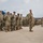 A formation of U.S. Army soldiers in Operational Camouflage Pattern (OCP) uniforms are standing at attention in a paved outdoor area, likely a parking lot or parade ground. A large blue bus is visible in the background, suggesting a deployment, mobilization, or unit assembly event. The soldiers are arranged in multiple rows extending into the background.