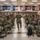 A large group of U.S. Army soldiers in OCP (Operational Camouflage Pattern) uniforms are seated in an auditorium or assembly hall, applauding as a fellow soldier walks down the center aisle. The room is decorated with numerous U.S. state flags hanging along the upper walls, suggesting a military installation with ceremonial significance. The setting appears to be a formal briefing, ceremony, or graduation event.