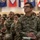 A group of U.S. Army soldiers are seated in an auditorium or briefing hall, attentively listening to a presentation or lecture. The soldier in the foreground, identified by the name tape 'USNER,' is wearing the Army Combat Uniform (ACU) in Operational Camouflage Pattern (OCP) and holds a small notebook. Multiple military unit flags are visible hanging in the background.