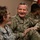 Two U.S. Army soldiers are engaged in a cheerful conversation, with the central figure laughing broadly. The soldier in the center wears a name tape reading 'BERGER' and displays a two-star rank insignia, indicating the rank of Major General. Additional soldiers are visible seated in the background in a casual indoor setting.