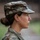 A profile portrait of a female U.S. Army soldier wearing Operational Camouflage Pattern (OCP) uniform and patrol cap, photographed in rainy conditions. She is gazing forward with a focused expression. A U.S. flag patch with gold border is visible on her left shoulder, and a partial name tape reading 'KUCK' (or similar) is visible on her chest.