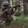 A U.S. Army soldier in full combat gear is kneeling in a wooded area while aiming an M16-style rifle equipped with a blank firing attachment (red cap on muzzle). At least one additional soldier is visible in the background in a low position. The scene appears to be a field training exercise based on the blank firing attachment and training environment.