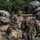 A U.S. Army soldier, identified by name tape as 'Underwood,' addresses a group of soldiers in full combat gear including helmets and tactical equipment. The central figure is in focus, wearing an OCP (Operational Camouflage Pattern) uniform with a patrol cap, and appears to be giving instructions or a briefing. The foreground soldiers are intentionally blurred, creating a depth-of-field effect that emphasizes the speaking soldier.