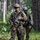 Two U.S. Army soldiers in full combat gear are patrolling through a forested area. Both are wearing MultiCam (OCP) camouflage uniforms, helmets, and carrying M4/M16-style rifles with training devices attached. The image appears to depict a field training exercise based on the MILES-style laser tag devices visible on the weapons.