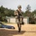 A soldier in full combat gear walks across a gravel area at what appears to be a military training facility. The soldier is carrying a rifle and an ammunition can while wearing OCP (Operational Camouflage Pattern) uniform, tactical vest, helmet, and protective eyewear. The background includes storage buildings, trees, and a civilian vehicle, consistent with a mock urban training environment.