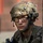 A young male soldier in full combat gear is positioned against a stone or concrete column, appearing to be in a tactical stance. He is wearing a multicam-patterned helmet with accessories attached, black-framed glasses, and a matching multicam uniform. He is holding what appears to be a firearm and is looking alertly to his left.