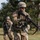 Two U.S. Army soldiers in full combat gear are moving across an open grassy field with trees in the background. The foreground soldier is in sharp focus, wearing MultiCam (OCP) camouflage, a combat helmet, tactical sunglasses, and carrying an M16/M4-style rifle with a blank firing attachment (BFA) on the muzzle. The setting and equipment suggest a field training exercise (FTX) rather than a combat deployment.