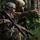 Two U.S. Army soldiers in full combat gear are positioned in a wooded area during what appears to be a field training exercise. One soldier is pointing in a direction while the other observes, suggesting coordination or target identification. Both are armed with M16/M4-style rifles and wearing OCP (Operational Camouflage Pattern) uniforms with tactical helmets.