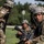 The image shows a group of U.S. Army soldiers in full combat gear conducting what appears to be a field training exercise. Multiple soldiers wearing ACU/OCP camouflage uniforms and combat helmets are visible, with at least one carrying an M16/M4-style rifle. The setting is an outdoor grassy area with trees in the background, suggesting a training base or exercise area.
