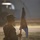 A silhouetted U.S. Army soldier in combat uniform stands at attention holding a military guidon or unit flag during what appears to be a formal military ceremony or formation. The image is backlit by low-angle sunlight, creating a dramatic silhouette effect. A large military vehicle or aircraft is visible in the blurred background.