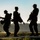 Three military personnel in camouflage uniforms are silhouetted against an early morning or late evening sky, walking across what appears to be a training or base area. They are carrying tactical gear, backpacks, and equipment consistent with a field exercise or ruck march. The backlighting creates a strong silhouette effect with a misty treeline visible in the background.