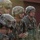 A group of military personnel in full combat gear are standing in formation, appearing to receive a briefing or instructions. They are wearing ACU (Army Combat Uniform) and OCP (Operational Camouflage Pattern) uniforms with tactical helmets and equipment. The image is captured in warm, backlit lighting suggesting early morning or late afternoon conditions.