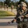A U.S. Army soldier in the foreground carries an M16-style rifle with a training device (MILES gear or blank firing adapter) attached, wearing ACU/OCP camouflage uniform, helmet, and protective eyewear. The soldier is moving along a paved road during what appears to be a military training exercise. Multiple additional soldiers in similar gear are visible in the background, suggesting a unit-level field training exercise.