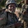 A U.S. Army soldier in full combat gear smiles while holding what appears to be a mortar round or similar cylindrical military equipment. The soldier is wearing MultiCam (OCP) pattern uniform with a U.S. flag patch on the left shoulder, a combat helmet with chin strap, and body armor. Additional soldiers in similar uniforms are visible in the blurred background.
