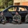 Three soldiers in US Army OCP (Operational Camouflage Pattern) uniforms are seated on the ground in front of a heavily damaged black sedan, appearing to be on a break during a training exercise. They are equipped with rifles and tactical gear, and appear relaxed and conversational. The setting appears to be a military training facility with a mock urban environment structure visible in the background.