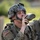 A U.S. Army soldier in full combat gear is drinking from a military-issue canteen. The soldier is wearing OCP (Operational Camouflage Pattern) uniform with an ACH (Advanced Combat Helmet) in UCP (Universal Camouflage Pattern) and carries a rucksack. A U.S. flag patch is visible on the left shoulder, indicating active-duty or reserve/National Guard status.