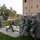 A group of military soldiers in full combat gear are conducting an urban warfare training exercise near a multi-story concrete block building. Soldiers are positioned along a wall in a tactical stack formation while one soldier moves through an opening, suggesting a building-clearing or breach drill. The setting appears to be a dedicated military training facility with constructed obstacle walls and a mock urban environment.