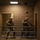 Two armed military personnel in full combat gear are positioned in what appears to be an indoor training facility or mock building. They are in tactical stances behind a metal railing on what looks like a second-floor corridor or balcony area. The setting and postures suggest a military training exercise involving building clearance or close-quarters combat drills.