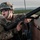 A U.S. Army soldier in full combat gear is positioned behind a concrete barrier, holding an M16-style rifle in a tactical firing position. The soldier is wearing MultiCam/OCP camouflage, an ACH helmet, and protective eyewear, with mouth open as if shouting a command. The background shows a forested, hilly landscape under an overcast sky, suggesting a training exercise environment.