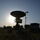 The image shows a military or communications field installation photographed in silhouette against a low sun, creating strong backlit conditions. Multiple satellite dish antennas and ground equipment are deployed across an open grassy field. The scene appears to be a temporary or field-deployed communications or radar station.