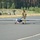 A military service member in camouflage uniform is manually positioning a fixed-wing unmanned aerial vehicle (UAV) on a paved surface, likely a runway or taxiway. The UAV appears to be a RQ-7 Shadow or similar tactical reconnaissance drone used by the U.S. Army. The setting appears to be a military airfield surrounded by pine forest, with ground support equipment visible in the background.