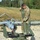 A U.S. Army soldier in MultiCam/OCP camouflage uniform is preparing or configuring a small unmanned aerial vehicle (UAV) on a launch rail system. The UAV appears to be an RQ-7 Shadow or similar tactical drone mounted on a catapult launcher. The setting appears to be a military training area or forward operating base with a gravel surface and forested background.