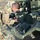 A U.S. Army soldier in Operational Camouflage Pattern (OCP) uniform is performing maintenance or diagnostic work on a piece of military equipment. The soldier is leaning over the equipment and appears to be using a handheld diagnostic or testing device. The equipment is olive drab green and bears a label indicating 'HYDRAULIC FLUID MIL-PRF-6083'.