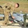 A military service member in multicam/OCP camouflage uniform is performing pre-flight or maintenance checks on a fixed-wing unmanned aerial vehicle (UAV), identified by the number '3276' on its fuselage. The UAV is mounted on a ground support launch or transport cradle on a gravel surface. The setting appears to be an open field training or operational area.