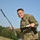 A smiling U.S. Army soldier in Operational Camouflage Pattern (OCP) uniform is posing outdoors while holding a military radio with an extended antenna in one hand and a clipboard or notepad in the other. He appears to be at a field training area with what appear to be unmanned aerial vehicles (UAVs/drones) visible on the ground behind him. The soldier displays a cheerful, relaxed demeanor suggesting a candid or informal photo moment.