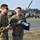 Two U.S. Army soldiers in OCP (Operational Camouflage Pattern) uniforms are standing on what appears to be a military installation or airfield. The soldier on the right is holding a radio controller with an extended antenna and a binder/clipboard, while the soldier on the left holds a handheld device. They appear to be engaged in a briefing or operational discussion.