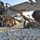 Two military personnel in US Army camouflage uniforms are performing maintenance or pre-flight inspection on what appears to be a fixed-wing unmanned aerial vehicle (UAV), likely a Shadow RQ-7 or similar tactical drone. The soldiers are lying on a gravel surface to access the underside of the aircraft. The image is taken from ground level, providing a close-up perspective of the aircraft's undercarriage and fuselage components.
