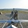 A military service member in camouflage uniform is performing pre-flight checks or maintenance on a fixed-wing unmanned aerial vehicle (UAV), likely an RQ-7 Shadow, positioned on a catapult launch rail system. The UAV is mounted on a pneumatic or hydraulic launch rail trailer on a gravel surface, with rolling hills and a paved runway visible in the background. The image appears to be taken at a forward operating base or military training area.