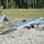 A U.S. Army soldier in MultiCam/OCP camouflage uniform is inspecting or preparing a fixed-wing unmanned aerial vehicle (UAV) marked with the number '3276', consistent with an RQ-7 Shadow or similar tactical UAS. The aircraft is positioned on a gravel surface with a forested background, suggesting a forward operating base or field training environment. The soldier appears to be communicating via radio while leaning over the UAV's tail section.