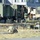 A military field operations setup showing multiple tactical vehicles and communications equipment deployed in an open area near a building. One soldier in camouflage uniform and headset is visible standing in the doorway of a shelter unit mounted on a military truck. The scene appears to be a command post or communications node establishment.