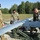 Three military personnel in MultiCam/OCP camouflage uniforms are preparing or inspecting a fixed-wing unmanned aerial vehicle (UAV) on a gravel surface. The drone, marked with the number '3276', is mounted on a green military launch or transport cradle. The setting appears to be an outdoor field area with trees visible in the background.