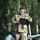 A U.S. Army soldier wearing OCP (Operational Camouflage Pattern) uniform is walking across what appears to be a tarmac or airfield surface while reviewing documents on a clipboard. The soldier is equipped with aviation-style hearing protection headset and is carrying a ground marshaling wand with a red flag. In the background, what appears to be a fixed-wing unmanned aerial vehicle (UAV) or small aircraft is visible.
