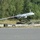 The image shows a fixed-wing unmanned aerial vehicle (UAV) marked with the number '3276' positioned on or near a catapult launch rail system on the ground. The drone appears to be a reconnaissance or surveillance type UAV with a pusher propeller configuration and sensor pod mounted beneath the fuselage. The setting appears to be a military airfield or test facility with a paved runway and tree line in the background.
