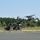 The image shows a military UAV (unmanned aerial vehicle) mounted on a catapult launch system attached to a wheeled military vehicle. The UAV appears to be in the process of being launched from the rail-based catapult system. The setting is an outdoor area near a paved road with dense tree line in the background.