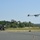 A fixed-wing unmanned aerial vehicle (UAV) is captured moments after launch from a catapult launch system mounted on a wheeled military vehicle. The UAV is airborne just above the runway surface with landing gear still deployed. The scene appears to be a military airfield or test facility surrounded by dense tree lines.